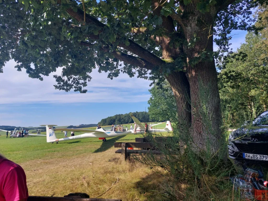 Mehrere Segelflugzeuge stehen auf dem Flugplatz des SFG Wershofen e.V., während Kinder und Erwachsene unter einem Baum im Vordergrund die Umgebung genießen.