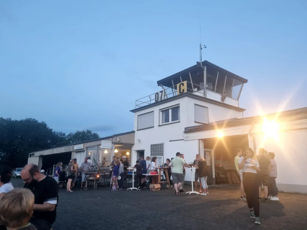 Menschen versammeln sich am Abend vor dem Gebäude des Flugplatzes des SFG Wershofen e.V., um das Sommerfest zu feiern.